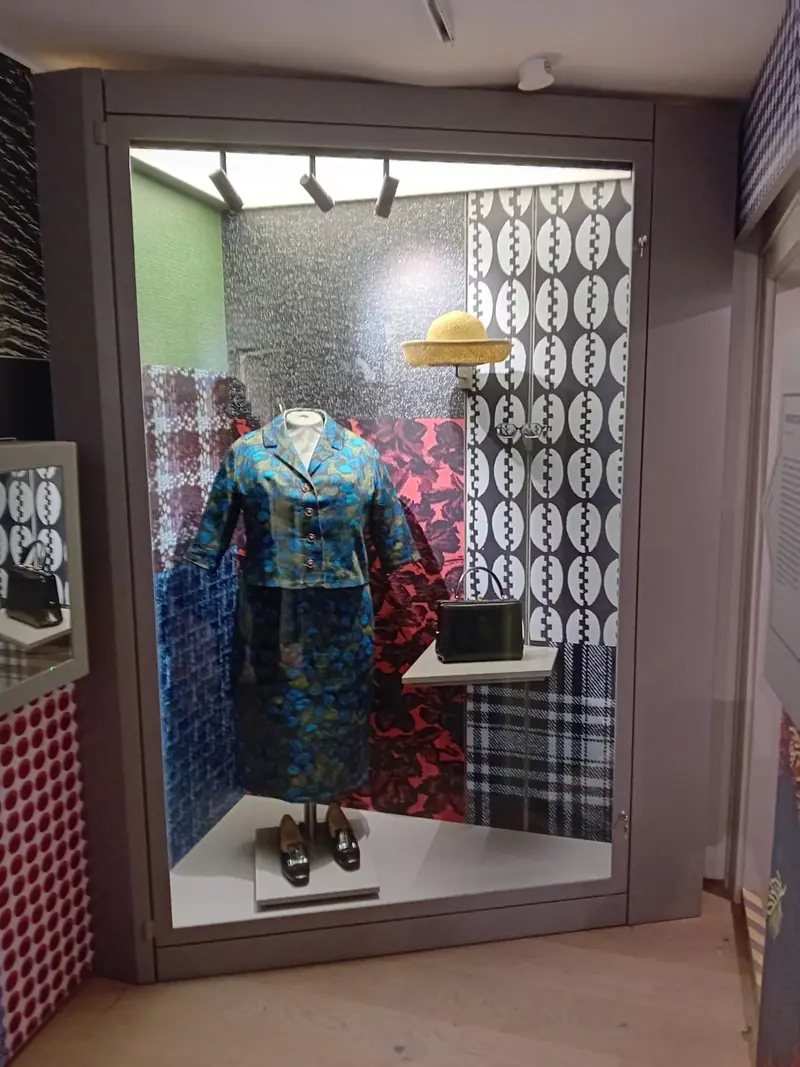 Beleuchtete Glasvitrine mit einer Figur in Kleidung vor gemustertem Hintergrund